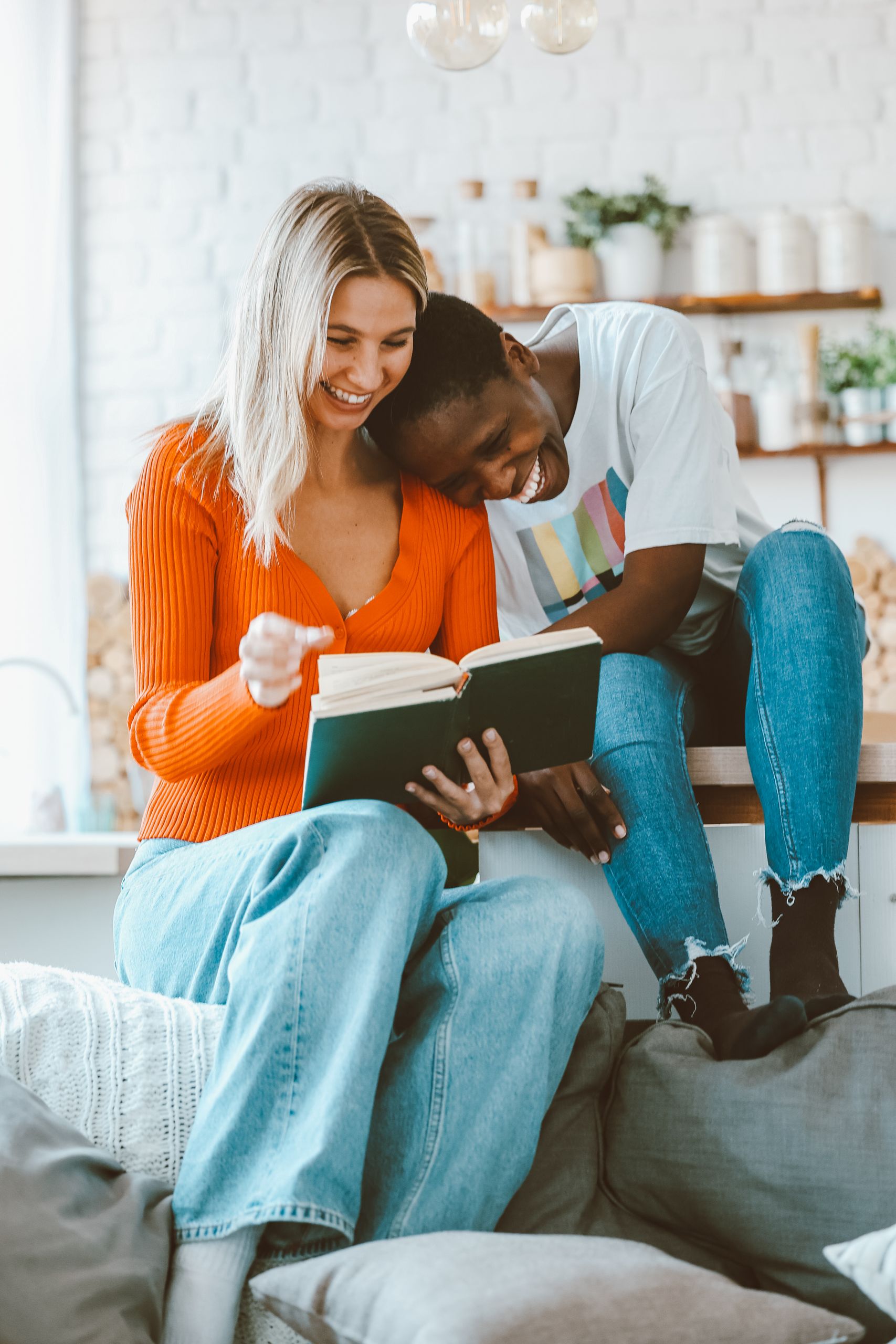 Two happy friends sitting on a couch, reading a book and enjoying each other's company at home.